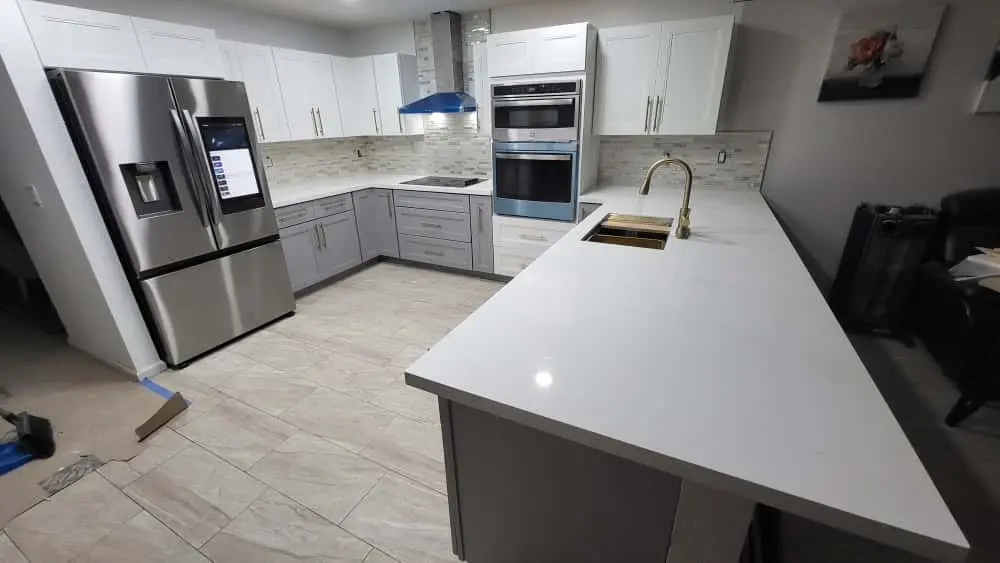 Completed kitchen with white shaker uppers, gray base cabinetry, a polished white peninsula, and brass sink and faucet
